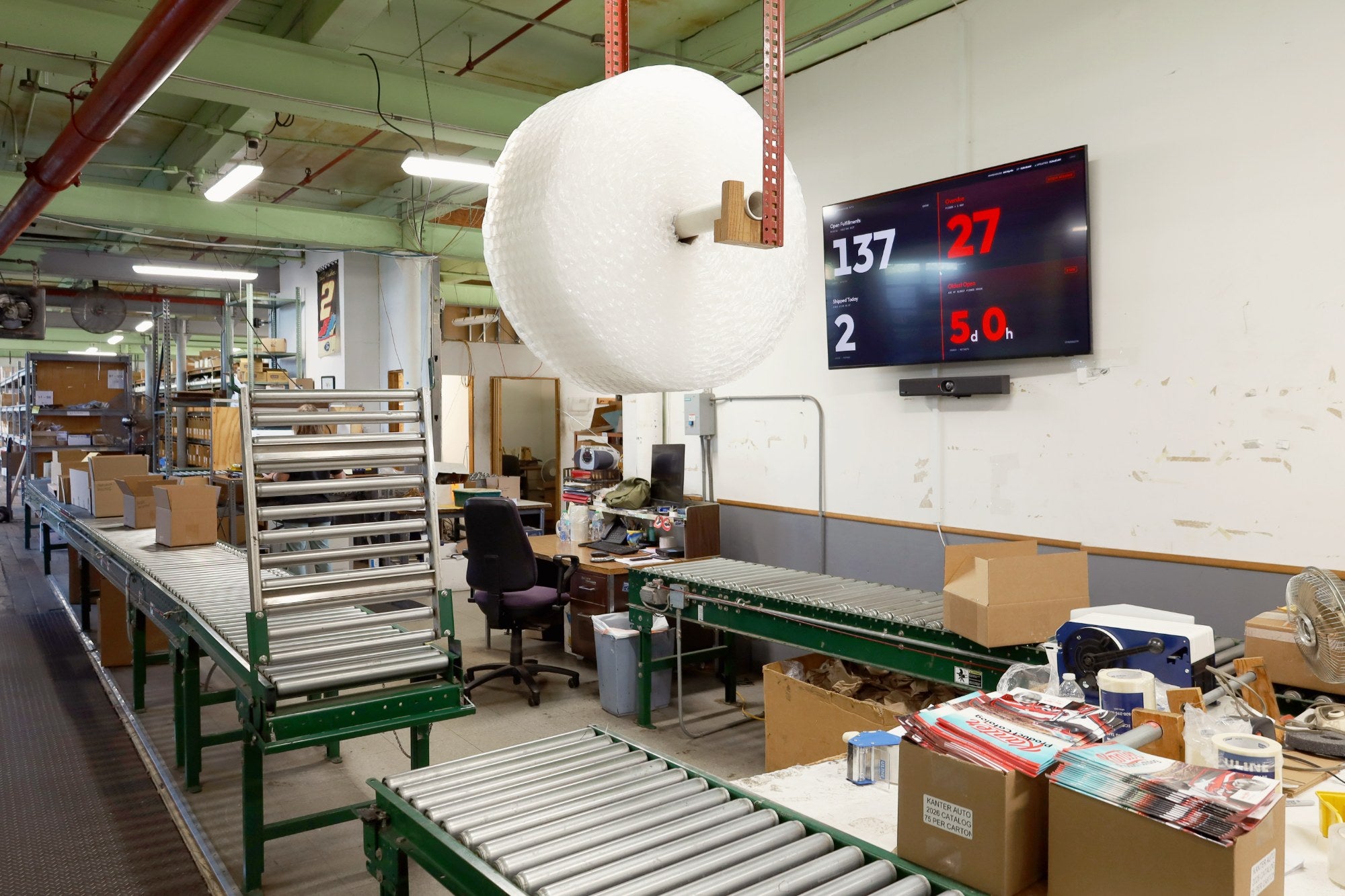 The Kanter warehouse floor with conveyors, a live fulfillment dashboard, and 2026 catalog boxes